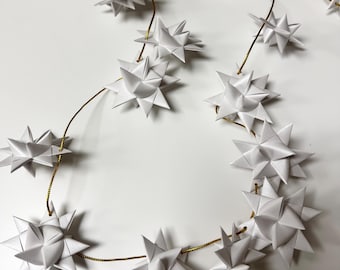 May include: A garland of white paper stars strung on gold wire. The stars have multiple points and are arranged in a flowing, decorative pattern. The background is a plain white surface.