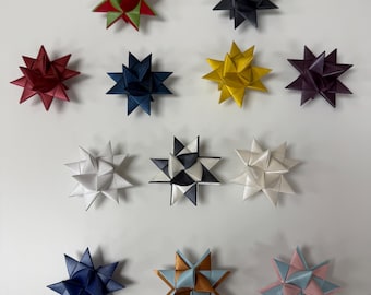 May include: A collection of origami star ornaments in various colors, including red, green, navy, yellow, and white. The stars are arranged on a white surface, showcasing their intricate folded designs. These decorative stars are ideal for holiday decor.