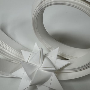 May include: A white paper star with pointed edges, positioned near a swirl of white paper strips. The star is the focal point, with the paper strips creating a dynamic background. The overall composition is clean and minimalist.