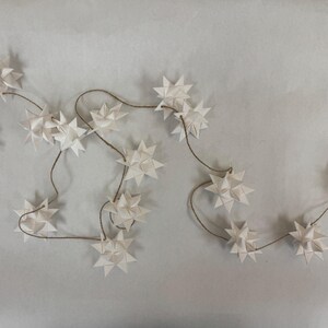May include: A string of white paper star ornaments on a brown string. The star-shaped decorations are evenly spaced along the string, creating a festive and decorative garland. The background is a light gray color.