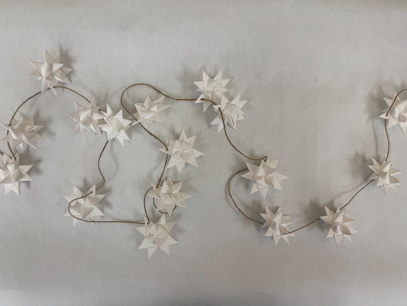 May include: A string of white paper star ornaments on a brown string. The star-shaped decorations are evenly spaced along the string, creating a festive and decorative garland. The background is a light gray color.
