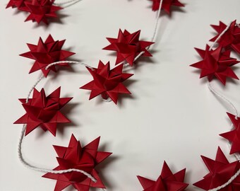 May include: A garland of red paper stars strung on white twine. The stars are multi-pointed and have a three-dimensional appearance. The garland is arranged in a curved pattern against a white background, suitable for holiday or festive decor.