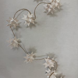 May include: A string of white paper star ornaments, each with multiple points, connected by a natural twine. The stars are arranged in a curved line against a light gray background, creating a festive and decorative display.