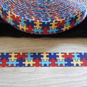 Może przedstawiać: Rolka wstążki w kolorze niebieskim, czerwonym, ż&oacute;łtym i białym z wzorem puzzli. Wstążka leży na czarnej powierzchni i drewnianej powierzchni.