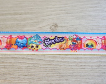 Może przedstawiać: Kolorowa wstążka z powtarzającym się wzorem postaci Shopkins. Wstążka przedstawia różne postaci Shopkins, w tym babeczkę, szminkę, rolkę sushi i truskawkę. Wstążka idealnie nadaje się do dodania odrobiny zabawy do każdego projektu rękodzielniczego.