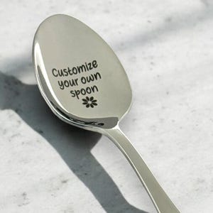 May include: A silver spoon with the text "Customize your own spoon" and a small flower design engraved on the front.