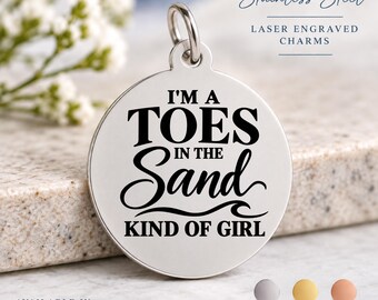 May include: A round stainless steel charm with a silver finish and a jump ring at the top. The charm features black laser-engraved text that reads I'M A TOES IN THE SAND KIND OF GIRL. The design includes a wave graphic below the word Sand.