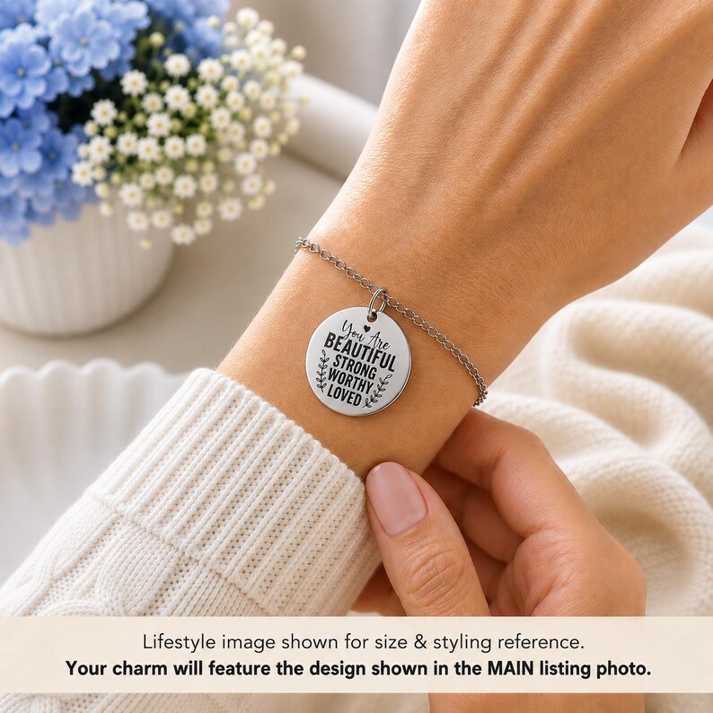 Op de afbeelding: Een zilverkleurige metalen kettingarmband met een ronde, platte bedel. De bedel is gegraveerd met de zwarte tekst: You Are BEAUTIFUL STRONG WORTHY LOVED, vergezeld van een klein hartsymbool en decoratieve bladaccenten aan de zijkanten.