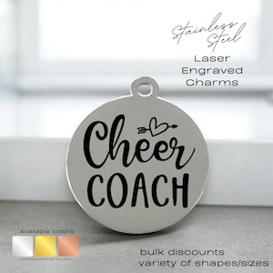 May include: A silver round charm with the text "Cheer COACH" in black lettering. The charm has a small arrow and heart design above the text. The charm is available in silver, gold, and copper.