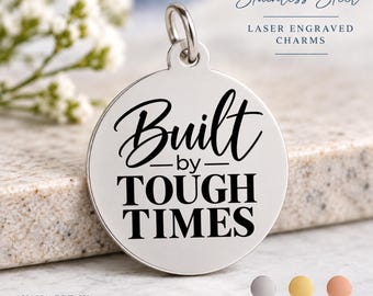 May include: A round stainless steel charm with a silver finish featuring the laser-engraved black text Built by TOUGH TIMES. The charm includes a jump ring at the top. Text on the image indicates Stainless Steel Laser Engraved Charms.