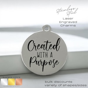 May include: Silver charm with the text "Created with a Purpose" engraved on it. The charm is round and has a loop at the top for attaching to a necklace or bracelet.