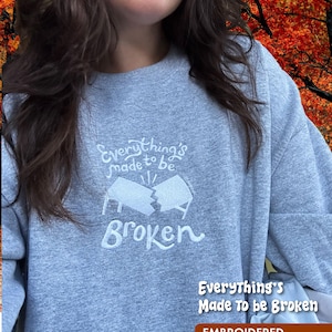 May include: A gray crewneck sweatshirt with white embroidered text that reads "Everything's made to be Broken." The design includes a broken flag graphic. The bottom right corner of the image has a brown rectangle with the words "EMBROIDERED CREWNECK".