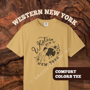 May include: Mustard-colored Comfort Colors tee with a Western New York graphic. The design features a person riding a buffalo, surrounded by floral elements, with the words "Western New York" in a vintage font. The shirt also has the text "Comfort Colors Tee".
