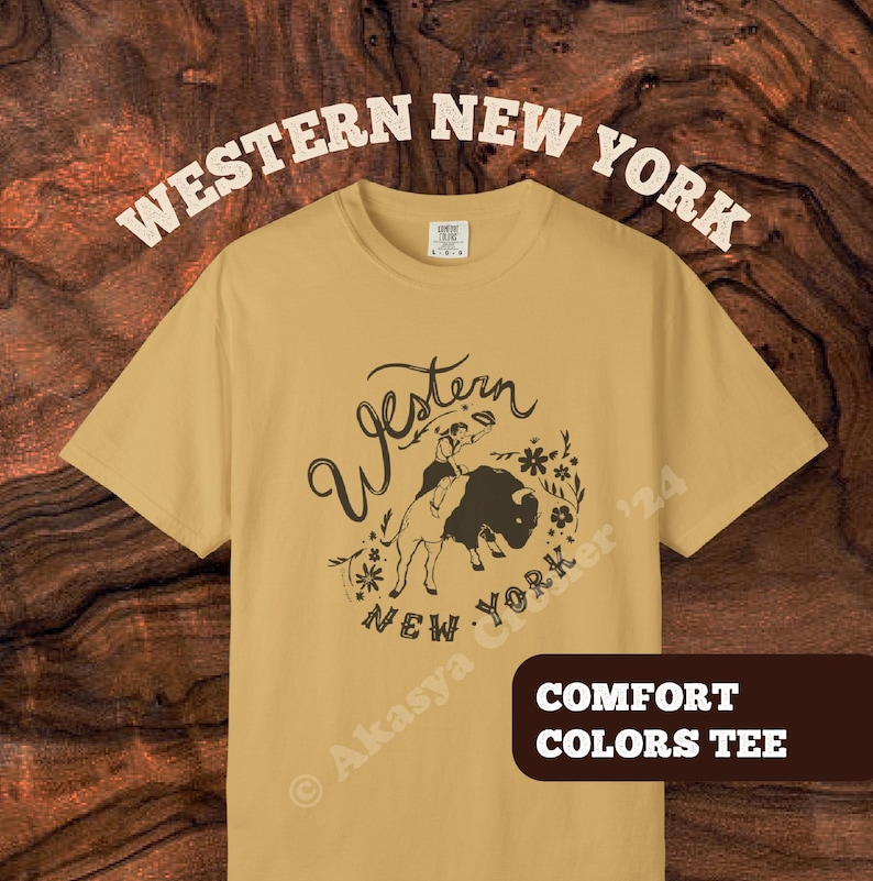 May include: Mustard-colored Comfort Colors tee with a Western New York graphic. The design features a person riding a buffalo, surrounded by floral elements, with the words "Western New York" in a vintage font. The shirt also has the text "Comfort Colors Tee".