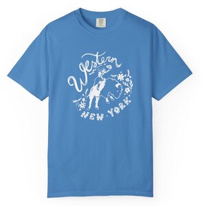 May include: A light blue t-shirt with a white graphic. The graphic features the words "Western New York" in a stylized font, along with an illustration of a person and a bull, surrounded by floral elements. The shirt has short sleeves and a crew neck.