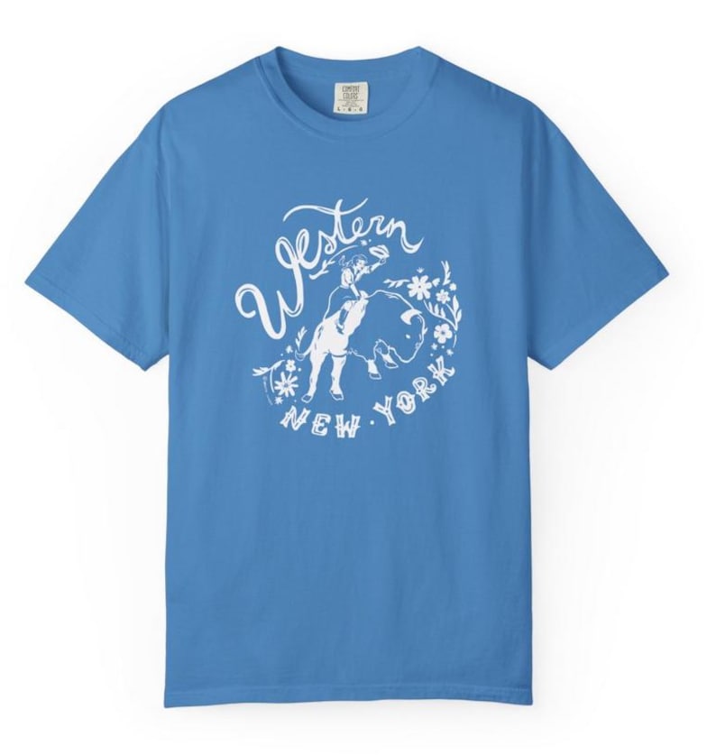 May include: A light blue t-shirt with a white graphic. The graphic features the words "Western New York" in a stylized font, along with an illustration of a person and a bull, surrounded by floral elements. The shirt has short sleeves and a crew neck.