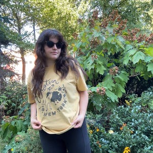 May include: Mustard yellow t-shirt with a graphic featuring a bison and the words "Western New York". The shirt is paired with black biker shorts. The image is taken outdoors with a background of green foliage.