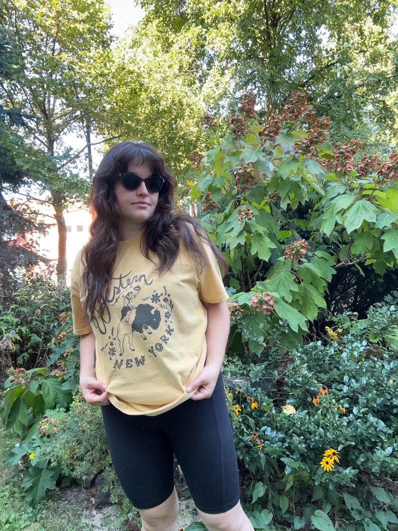 May include: Mustard yellow t-shirt with a graphic featuring a bison and the words "Western New York". The shirt is paired with black biker shorts. The image is taken outdoors with a background of green foliage.