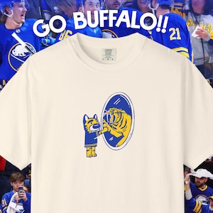 May include: Off-white t-shirt featuring a cartoon tiger looking in a mirror at itself. The mirror shows a tiger in a blue and gold uniform. The text "GO BUFFALO!!" is above the shirt, with a crowd of hockey fans in the background.