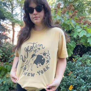 May include: Mustard yellow t-shirt with a graphic featuring a cowboy riding a bison, surrounded by floral elements, and the text "Western New York". The shirt is short-sleeved and made of a soft material.