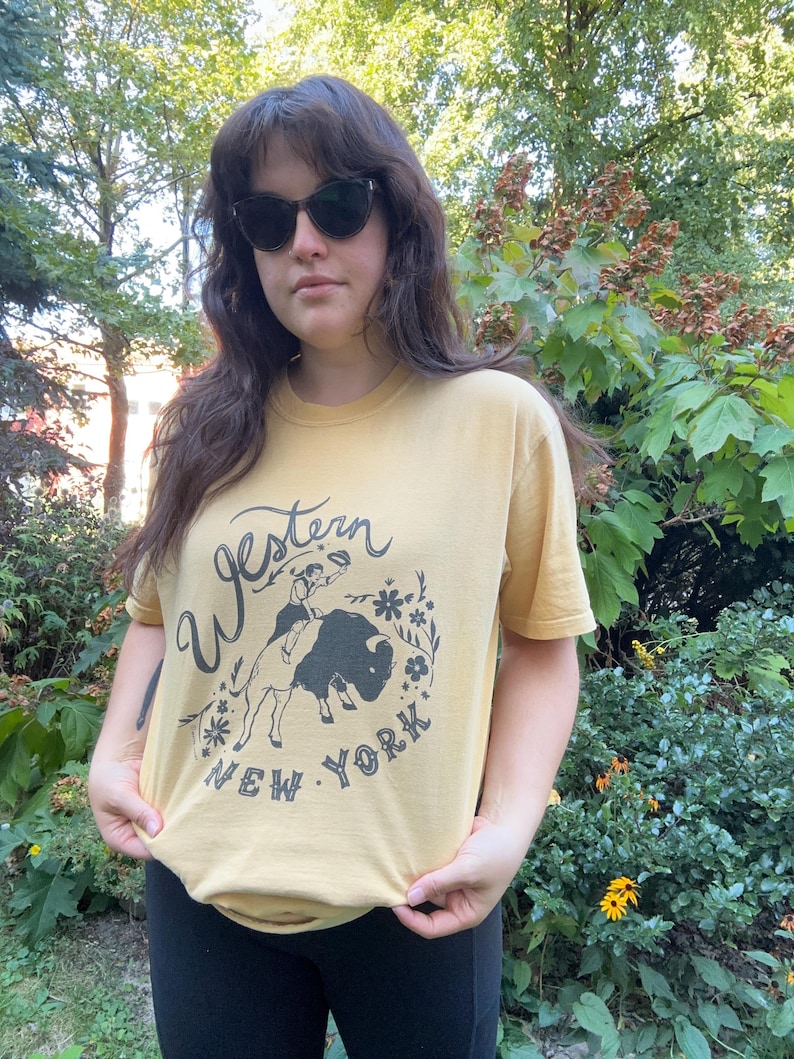 May include: Mustard yellow t-shirt with a graphic featuring a cowboy riding a bison, surrounded by floral elements, and the text "Western New York". The shirt is short-sleeved and made of a soft material.