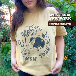 May include: Mustard yellow t-shirt with a black graphic of a person riding a bison, surrounded by floral elements. The text "Western New York" and "Comfort Colors Tee" are printed on the shirt.
