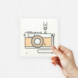 Op de afbeelding: Een zwart-witte lijntekening van een vintage camera met een geel en wit gepatroneerd lichaam. De camera heeft een band en een vinger drukt op de ontspanknop. De afbeelding is gesigneerd met "1/10" en "M.A."