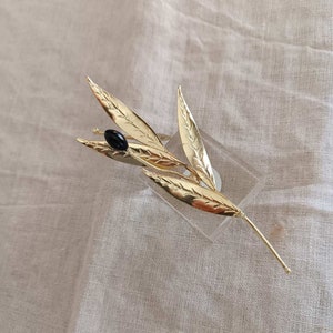 Op de afbeelding: Een goudkleurige bladbroche met gedetailleerde nerven en een enkele zwarte kraal die een olijf voorstelt. De broche wordt getoond op een heldere, rechthoekige standaard tegen een neutrale stoffen achtergrond. De broche is een decoratief accessoire.