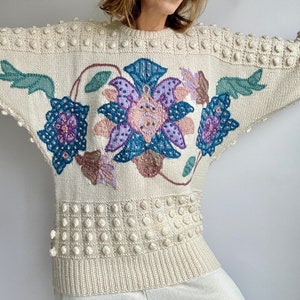 Puede incluir: Un suéter de punto color crema con un diseño floral en tonos de morado, azul y verde. El suéter tiene un diseño texturizado, con textura de bolitas.