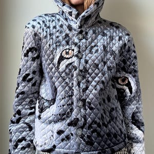 May include: A quilted jacket featuring a grey and black leopard print pattern with prominent feline eye graphics on the front panels. The garment has a high collar, long sleeves, and a vertical row of decorative silver-toned buttons down the centre.