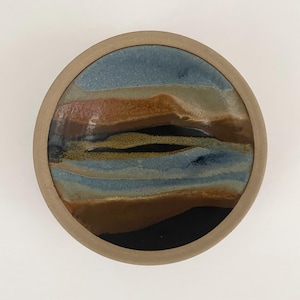 May include: A circular ceramic dish with a landscape design. The interior features layers of blue, brown, and black glazes, resembling a stylized landscape. The rim is a solid tan colour, providing a contrasting border. The dish is likely handmade.