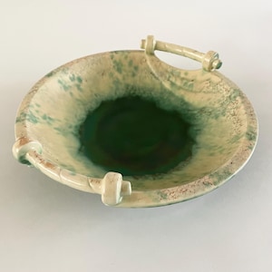 May include: A ceramic bowl with a handle. The bowl has a green interior and a cream-coloured exterior with green and brown speckles. It has a circular shape with a handle on top and two smaller handles on the sides.