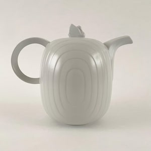 May include: A pale grey teapot with a rounded handle and a spout. The teapot has a lid with a small decorative feature. The body of the teapot has vertical, curved lines. The teapot is made of a solid, opaque material.