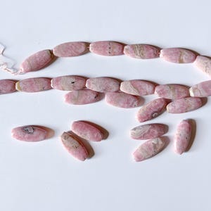 May include: A strand of pink oval-shaped beads with white and darker pink marbling. Several loose beads are scattered below the strand. The beads are strung on a thin, light pink string, suggesting jewelry making.