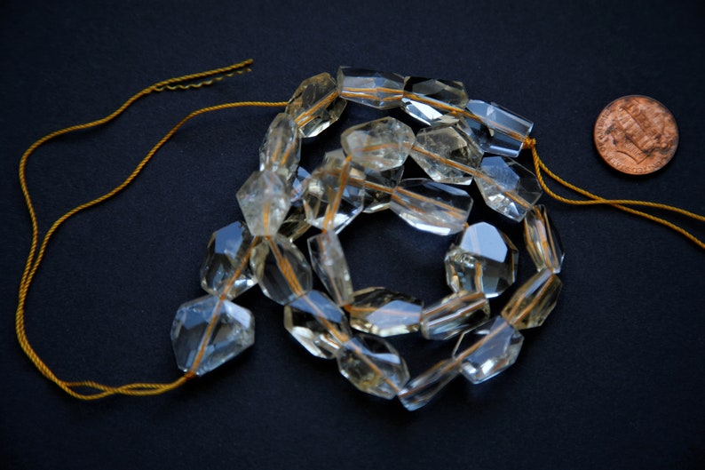 May include: A strand of clear quartz beads with a golden thread running through each bead. The beads are faceted and have a slightly yellow hue. The beads are strung on a gold cord.