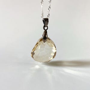 May include: A silver necklace with a pendant featuring a faceted, teardrop-shaped, light amber-coloured gemstone. The pendant is suspended from a delicate silver chain. The gemstone has a crystalline appearance, creating an elegant design.