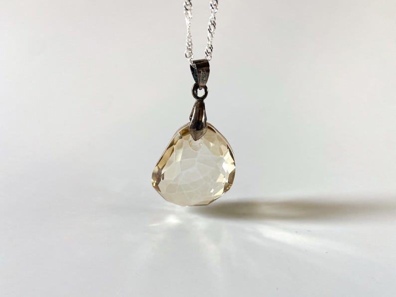 May include: A silver necklace with a pendant featuring a faceted, teardrop-shaped, light amber-coloured gemstone. The pendant is suspended from a delicate silver chain. The gemstone has a crystalline appearance, creating an elegant design.