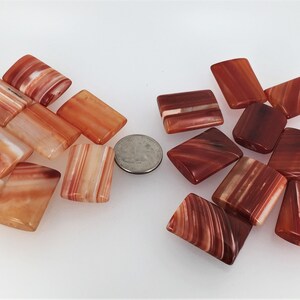 May include: A collection of carnelian gemstone beads, with a variety of red and orange hues, in a rectangular shape. The beads are arranged in two groups, with a US quarter coin for scale.