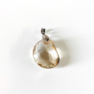 May include: A faceted, teardrop-shaped pendant with a light brown hue. The pendant is suspended from a small, silver-coloured bail. The gemstone is set against a white background.