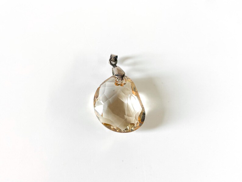 May include: A faceted, teardrop-shaped pendant with a light brown hue. The pendant is suspended from a small, silver-coloured bail. The gemstone is set against a white background.