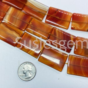May include: A strand of carnelian gemstone beads, each bead is a rectangular shape with a smooth, polished finish. The beads are a mix of orange and red hues with white banding. The beads are arranged in a line and are approximately 1 inch in length.