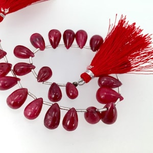 May include: A strand of red gemstone beads with a teardrop shape. The beads are strung on silver wire and have a red tassel at each end.