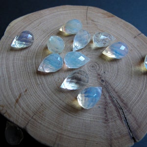 May include: A group of iridescent teardrop-shaped gemstones, with a milky white color, are arranged on a wooden surface.