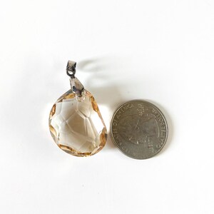 May include: A faceted, teardrop-shaped pendant with a silver-tone bail. The pendant is a light amber colour and is next to a coin for size comparison. The pendant has a geometric pattern.