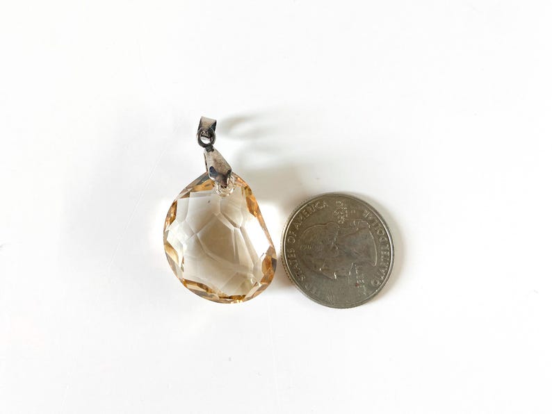May include: A faceted, teardrop-shaped pendant with a silver-tone bail. The pendant is a light amber colour and is next to a coin for size comparison. The pendant has a geometric pattern.