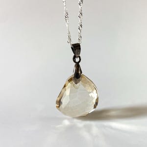 May include: A silver necklace featuring a faceted, teardrop-shaped, pale amber pendant. The pendant hangs from a delicate, twisted silver chain. The gemstone has a crystalline appearance.
