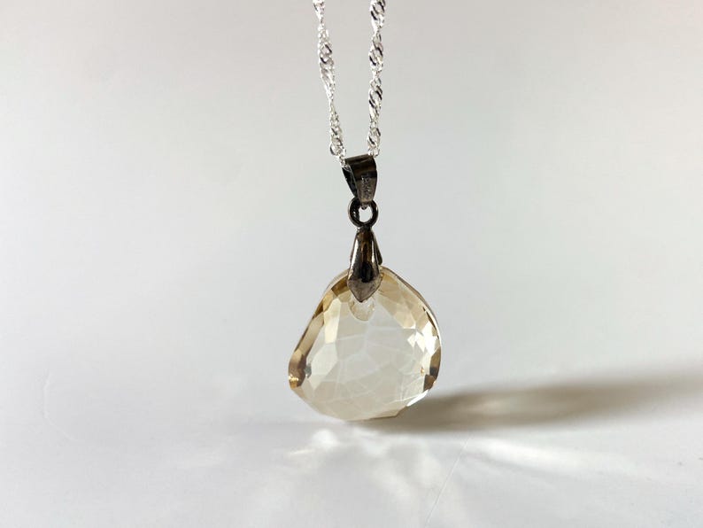May include: A silver necklace featuring a faceted, teardrop-shaped, pale amber pendant. The pendant hangs from a delicate, twisted silver chain. The gemstone has a crystalline appearance.