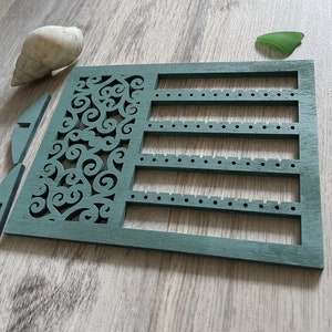 May include: A teal blue wooden earring holder with a decorative cutout design and rows of holes for storing earrings.