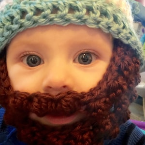 May include: A baby wearing a blue and white crocheted hat with a brown crocheted beard.