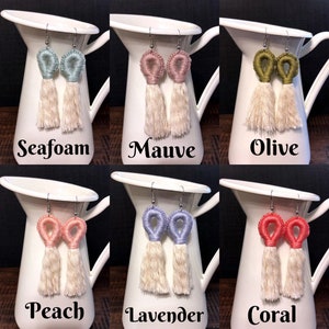 May include: Six pairs of handmade tassel earrings with different colored tops. The earrings are displayed on white ceramic pitchers. The colors are seafoam, mauve, olive, peach, lavender, and coral.